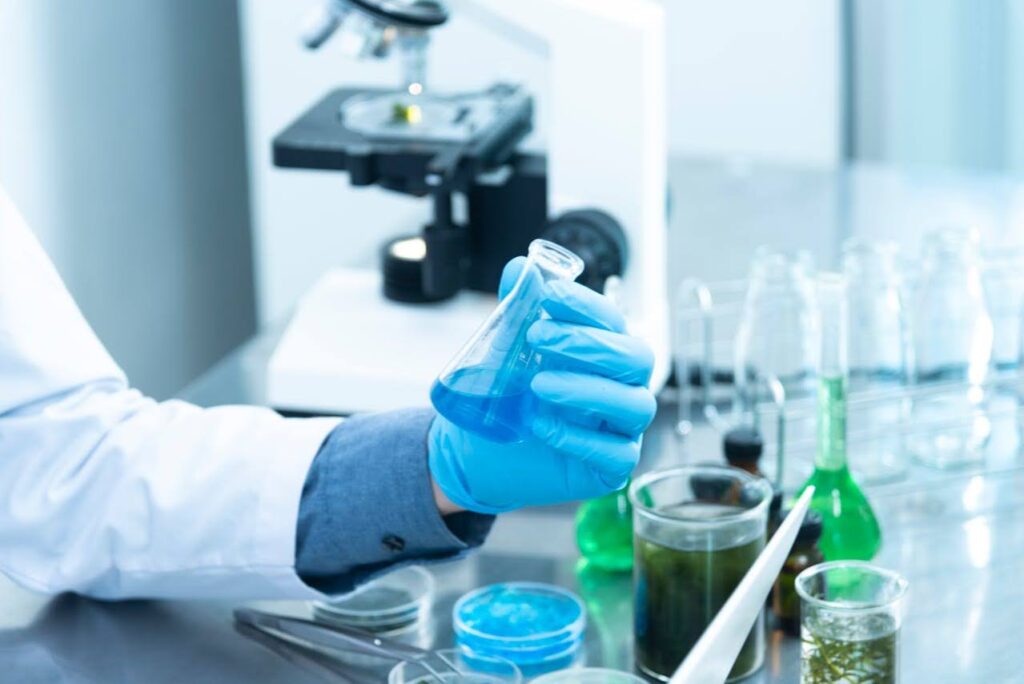 A person in a lab coat holds a test tube, surrounded by lab equipment and a microscope.