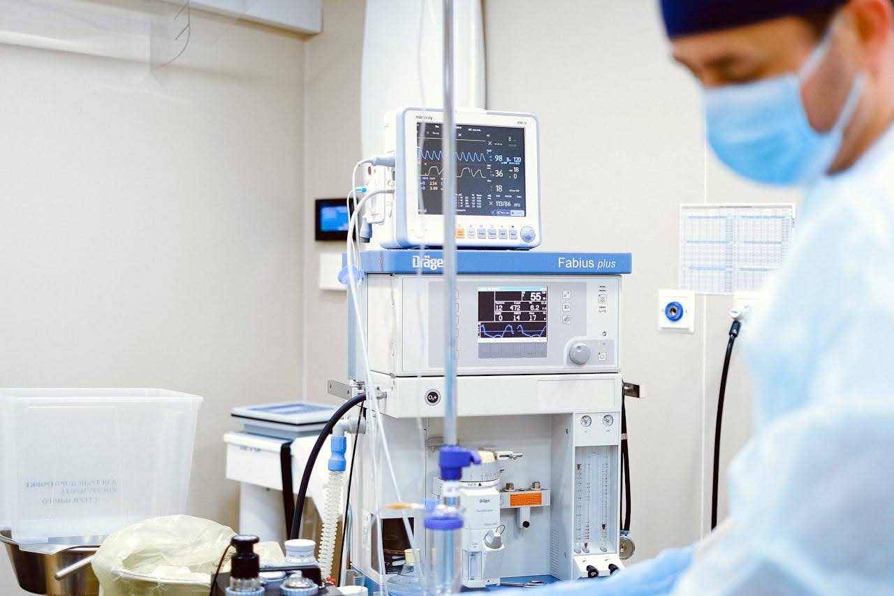 A doctor in a surgical mask stands near a machine in a hospital room, surrounded by medical equipment and monitoring screens.