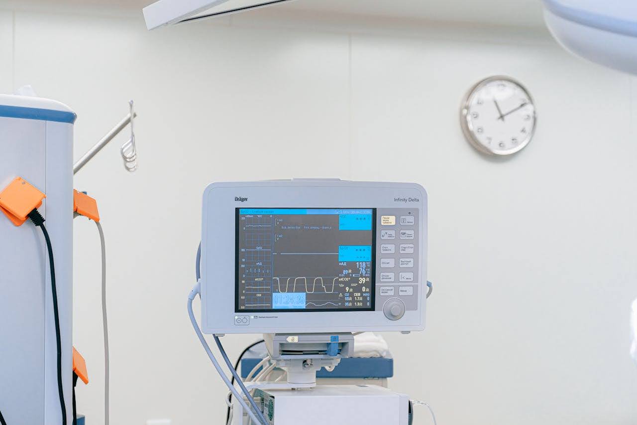 A monitor displays the patient's heart rate, blood pressure, oxygen levels, and vital signs, with medical equipment beside it and a clock in the background.