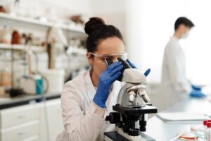 Biochemist wearing gloves and safety goggles examines samples using a microscope in a research laboratory.