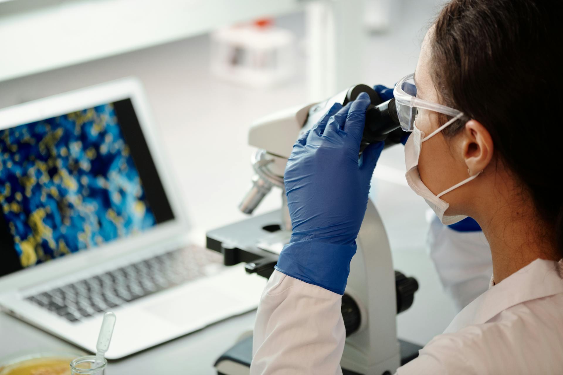 A young female scientist wearing protective gear examines examining samples through a microscope, with a laptop beside her displaying colourful cell imagery, in a precision medicine research lab.