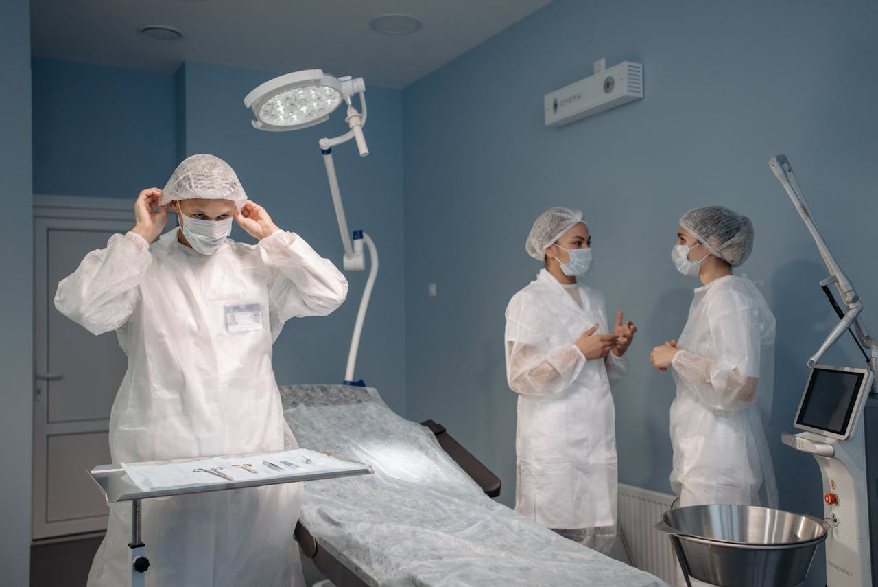 A general surgeon wearing a protective gown and surgical mask adjusts their cap in an operating room, while two medical staff members converse in the background.