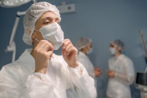 A close-up of a general surgeon putting on a surgical mask before a procedure, with two medical staff members blurred in the background.
