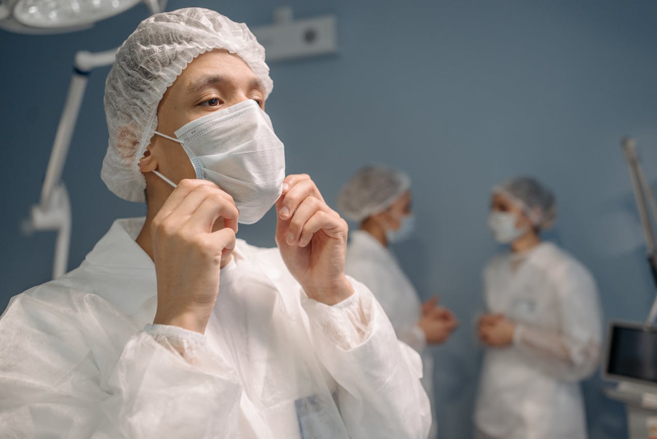 A close-up of a general surgeon putting on a surgical mask before a procedure, with two medical staff members blurred in the background.