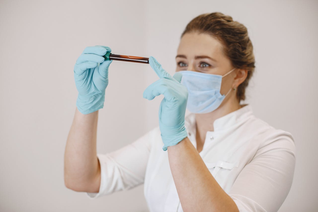 A female healthcare professional in gloves and a surgical mask holds a test tube filled with blood, examining it closely under bright lighting.