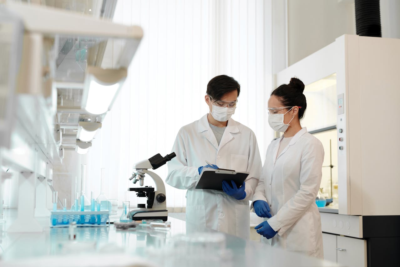 Two microbiologists in lab coats and masks discussing research findings beside a microscope in a modern laboratory.