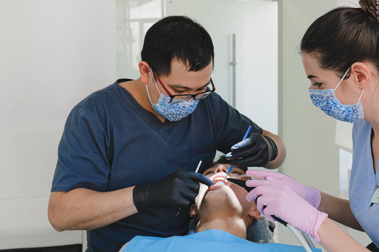 A male and female dental team performing a detailed dental procedure on a patient, both wearing patterned surgical masks and gloves, focused on patient care in a bright, sterile environment.