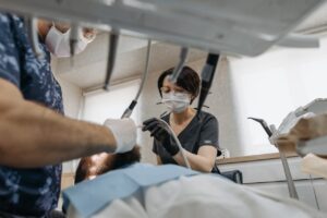 A female dentist wearing a surgical mask and gloves using dental instruments while treating a patient in a modern dental clinic, with another healthcare professional assisting.