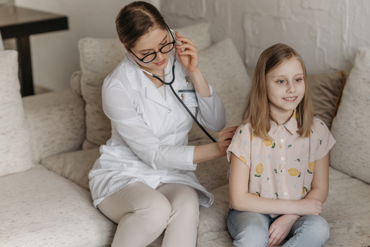 A female family medicine doctor using a stethoscope to examine a young smiling girl sitting on a couch during a home visit.