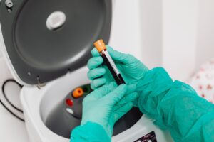 Gloved hands holding a blood sample vial near a centrifuge machine in a medical laboratory.