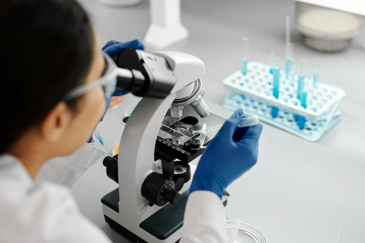 A female microbiologist examining a specimen slide through a microscope, with laboratory tools and test tubes visible in the background.