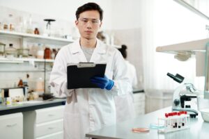 A young male microbiologist in a lab coat and safety goggles holding a clipboard in a laboratory setting with blood samples and a microscope on the bench.