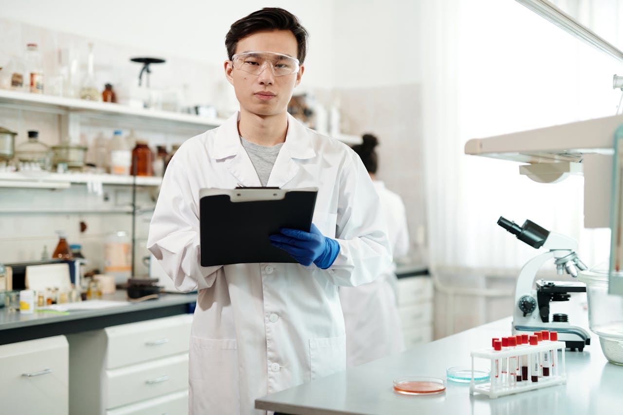 A young male microbiologist in a lab coat and safety goggles holding a clipboard in a laboratory setting with blood samples and a microscope on the bench.