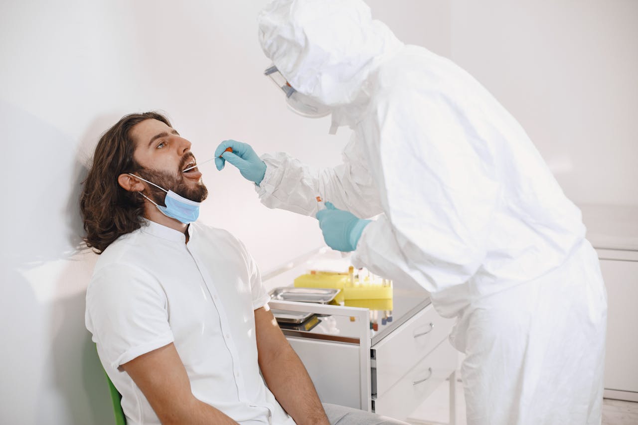An epidemiologist in full protective gear collects a swab sample from a seated patient during a medical test.