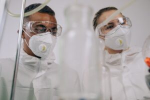 Two epidemiologists wearing protective masks and goggles examine samples through laboratory equipment in a research lab.
