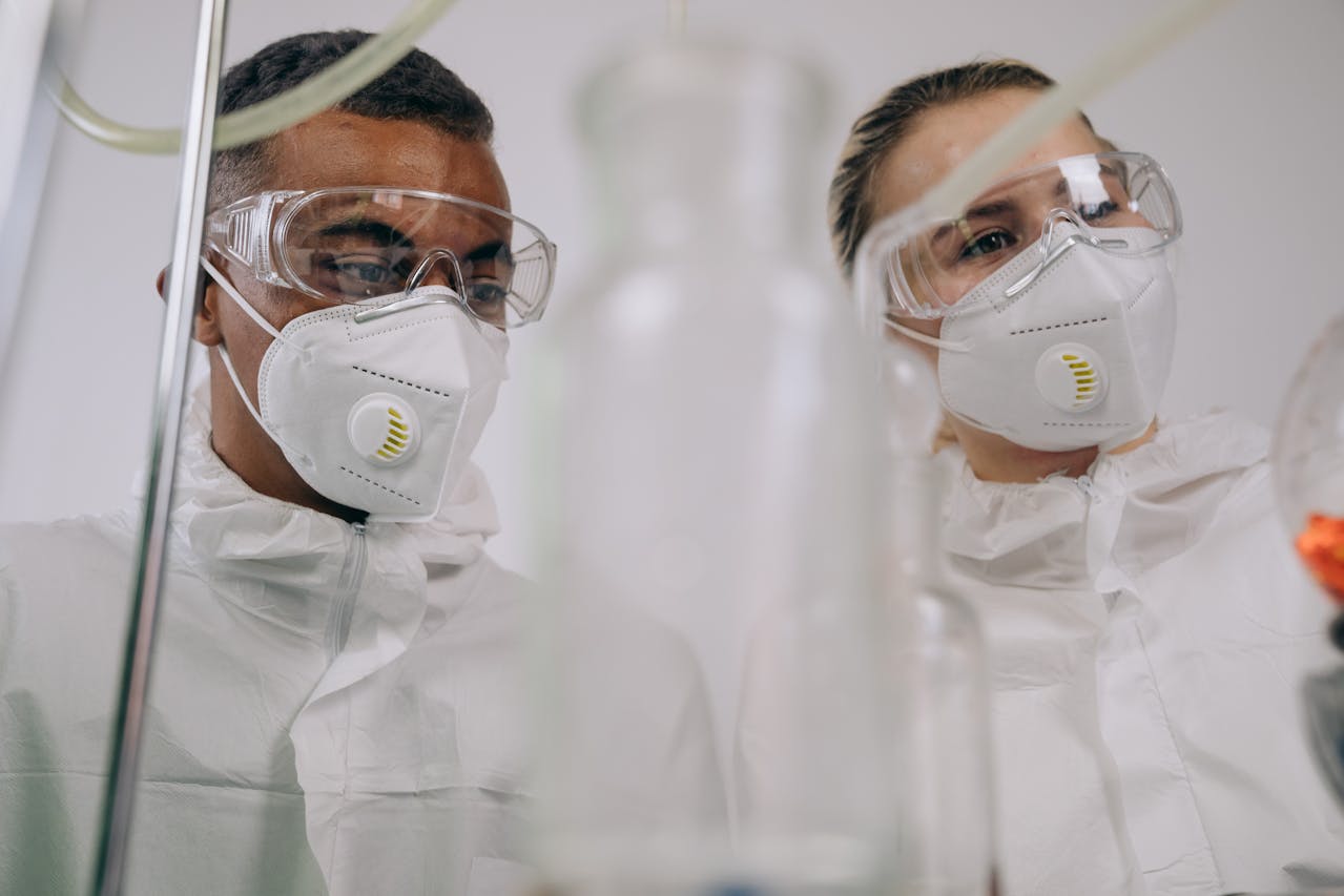 Two epidemiologists wearing protective masks and goggles examine samples through laboratory equipment in a research lab.