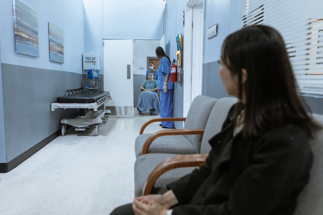 Patient sitting in a wheelchair in a hospital hallway attended by a nurse, with another person waiting on a bench.