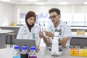 Two students in lab coats examine test tubes with colored liquids in a laboratory, with a microscope and other lab equipment on the table.