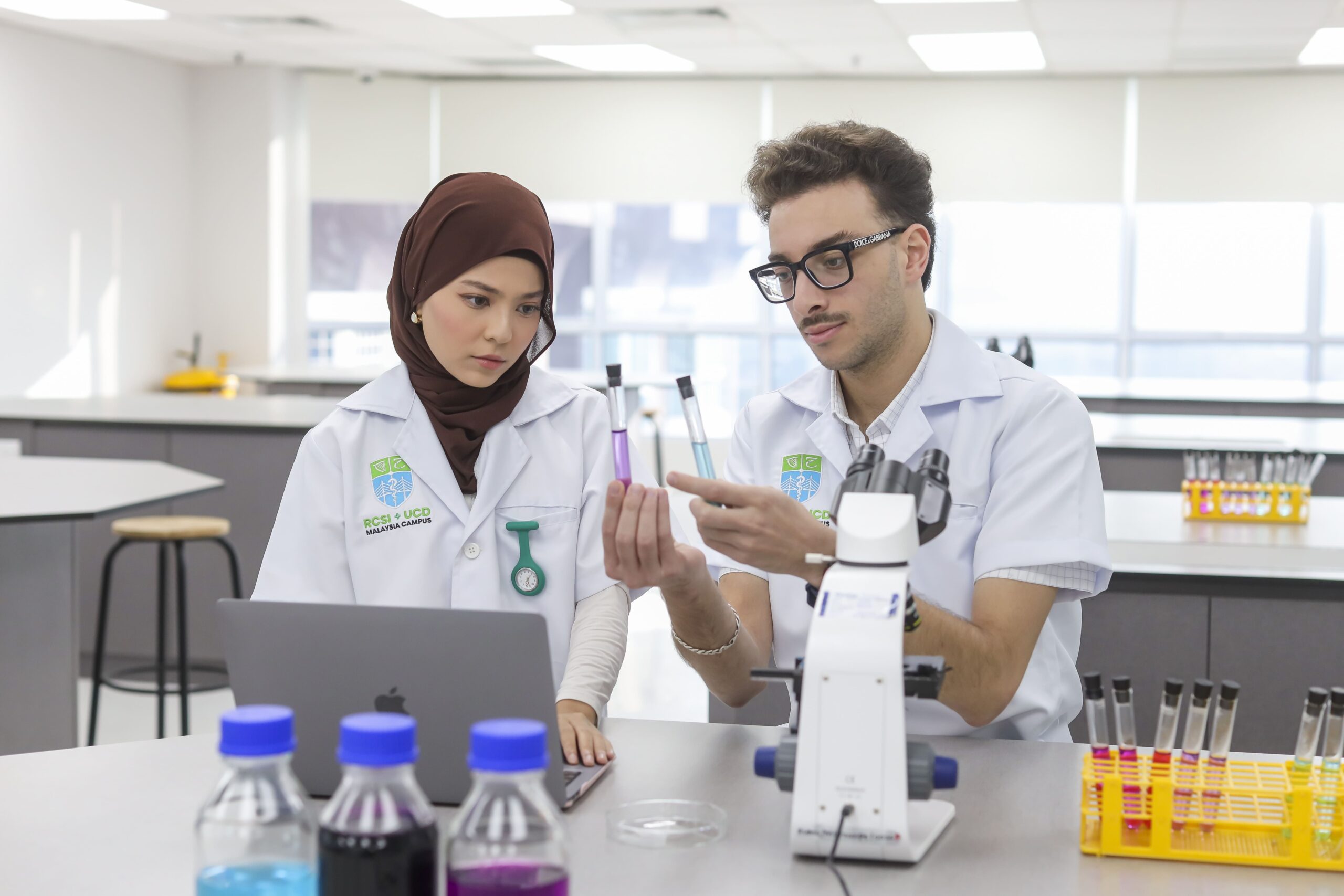Two students in lab coats examine test tubes with colored liquids in a laboratory, with a microscope and other lab equipment on the table.