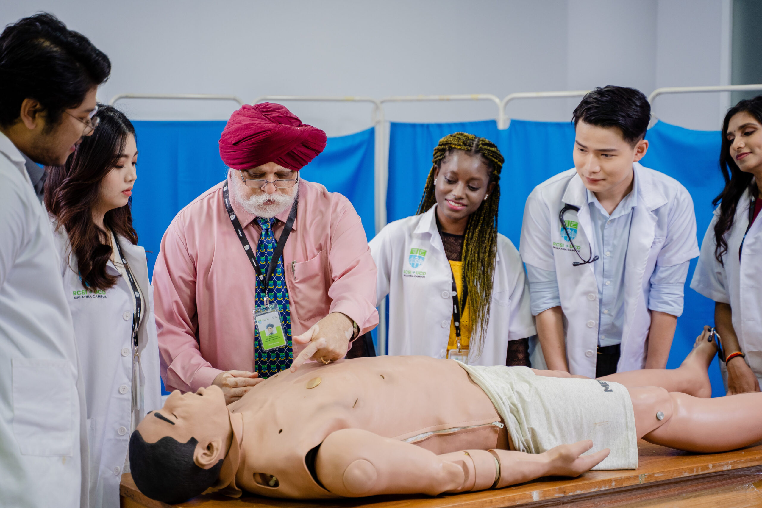 Instructor demonstrates an examination on a medical training mannequin while a group of medical students in lab coats stand around and observe in a clinical skills lab.