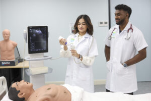 Two medical students in white lab coats practice using an ultrasound machine on a full-body training mannequin in a clinical skills room, while the screen shows an ultrasound image.