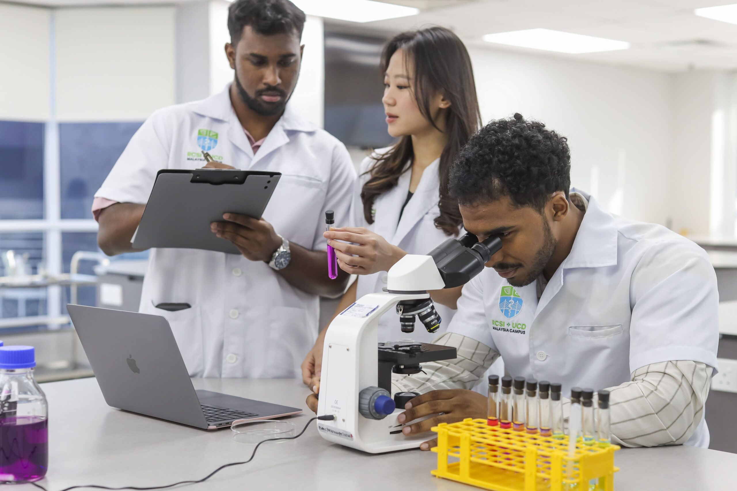 Three students in lab coats conduct a lab experiment, with one using a microscope, one holding a test tube, and another recording observations on a clipboard.