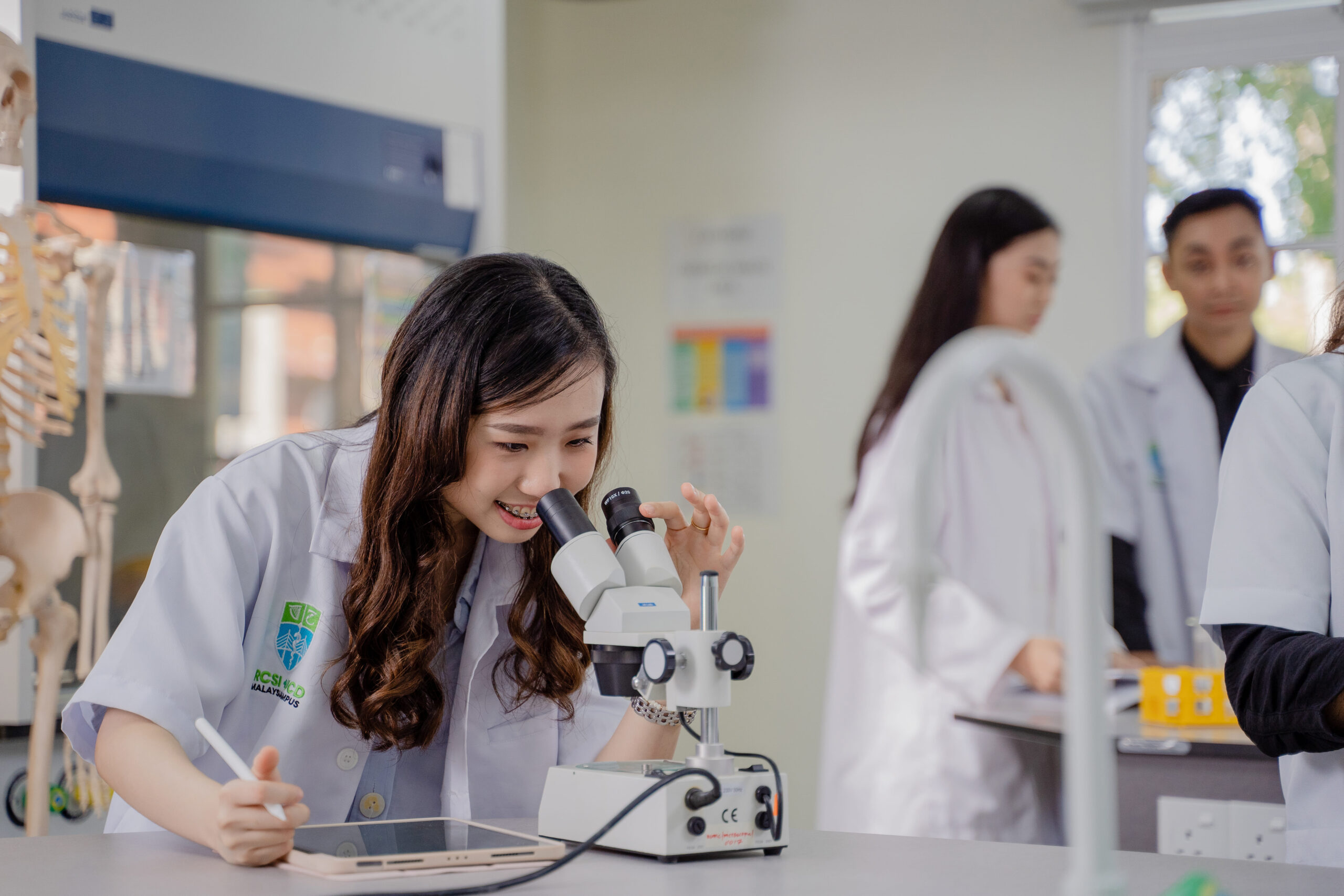 Medical student in a lab coat looks through a microscope while taking notes in a science lab, with other students working in the background.