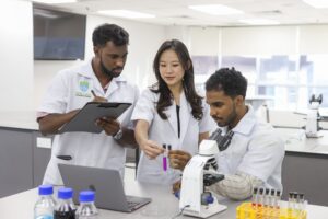 Three students in lab coats work together in a laboratory, examining test tubes while one takes notes and another uses a microscope.
