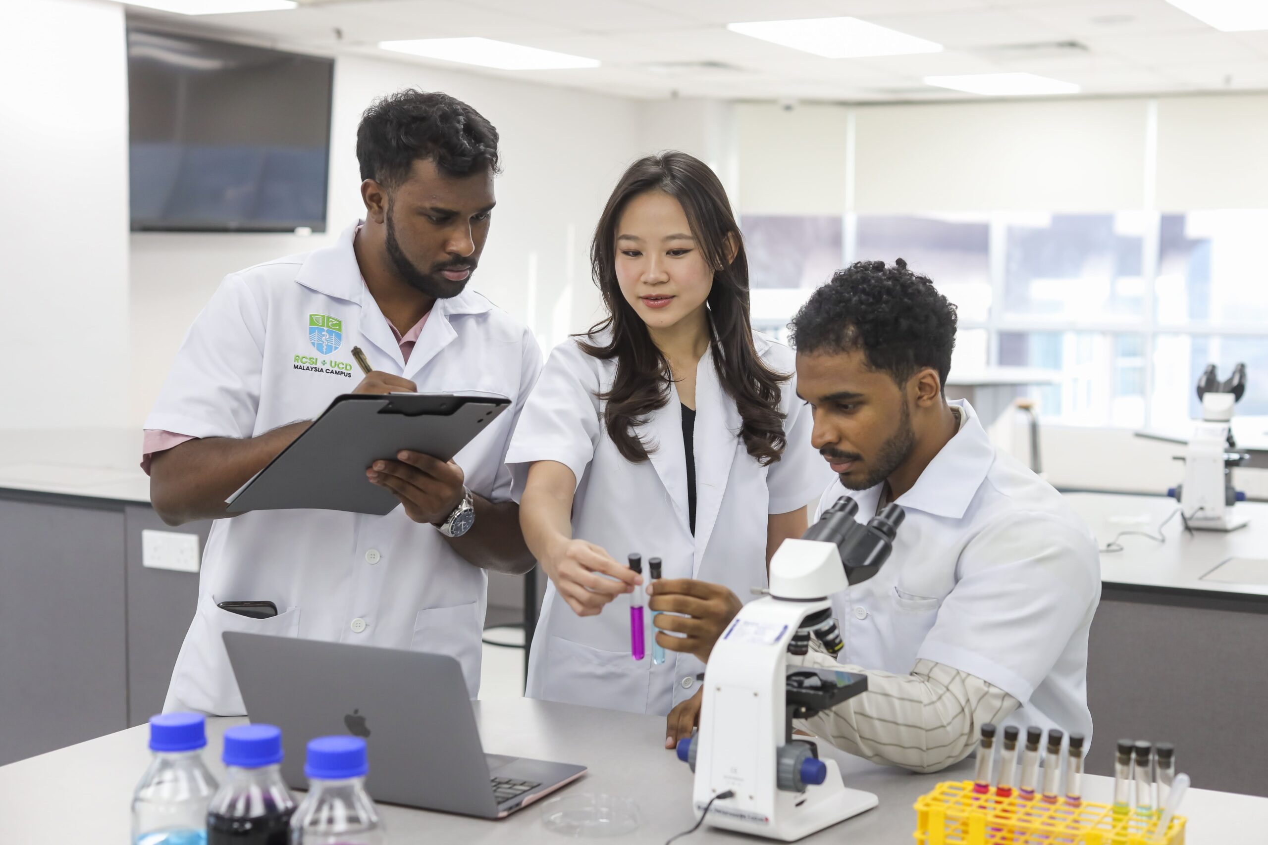 Three students in lab coats work together in a laboratory, examining test tubes while one takes notes and another uses a microscope.