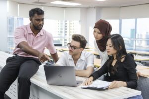 Four students collaborate around a laptop in a classroom, with one pointing at the screen while the others review notes and discuss.