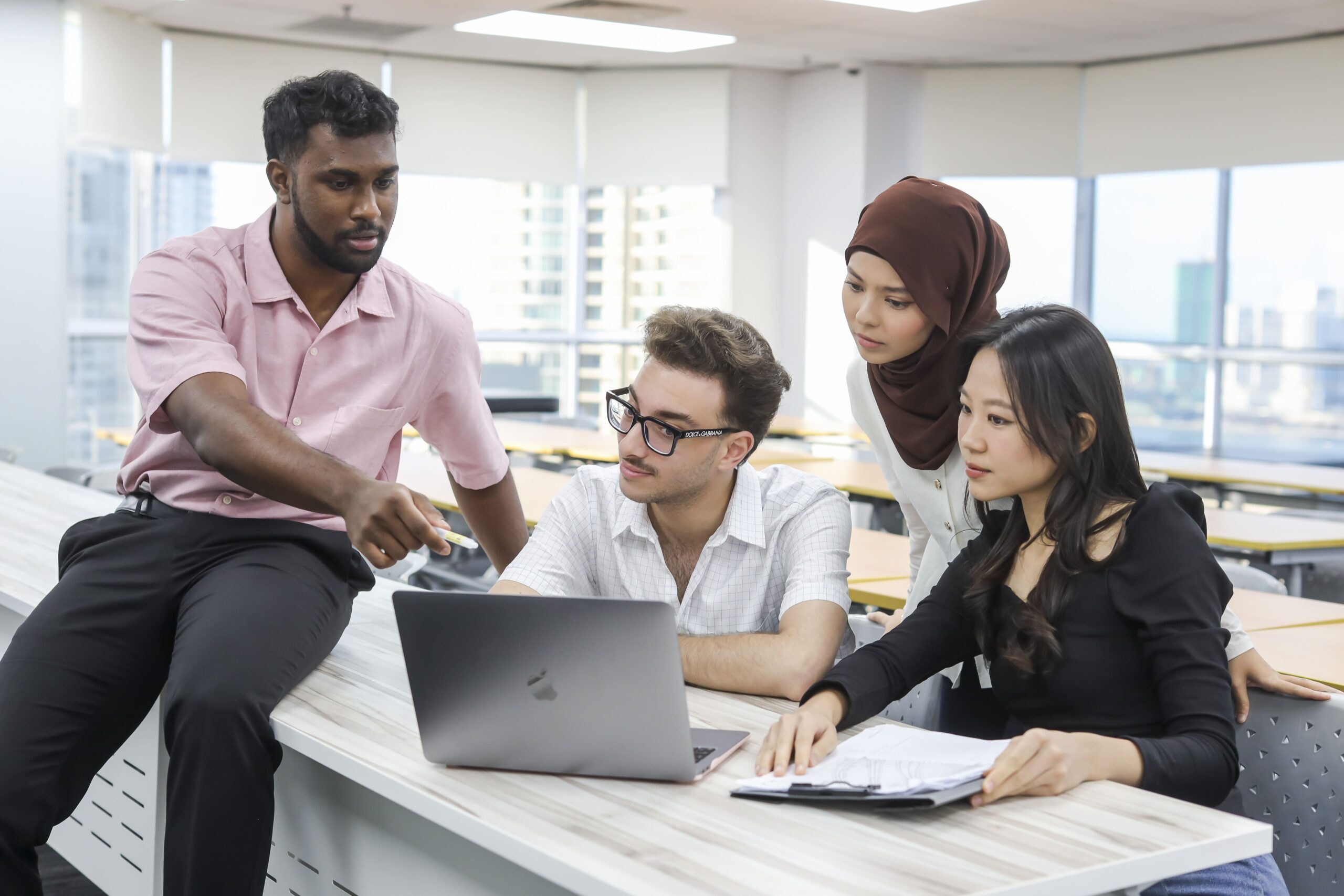 Four students collaborate around a laptop in a classroom, with one pointing at the screen while the others review notes and discuss.