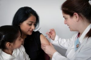 Public health nurse administering vaccine to woman with child observing.