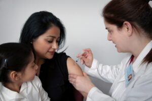 Public health nurse administering vaccine to woman with child observing.