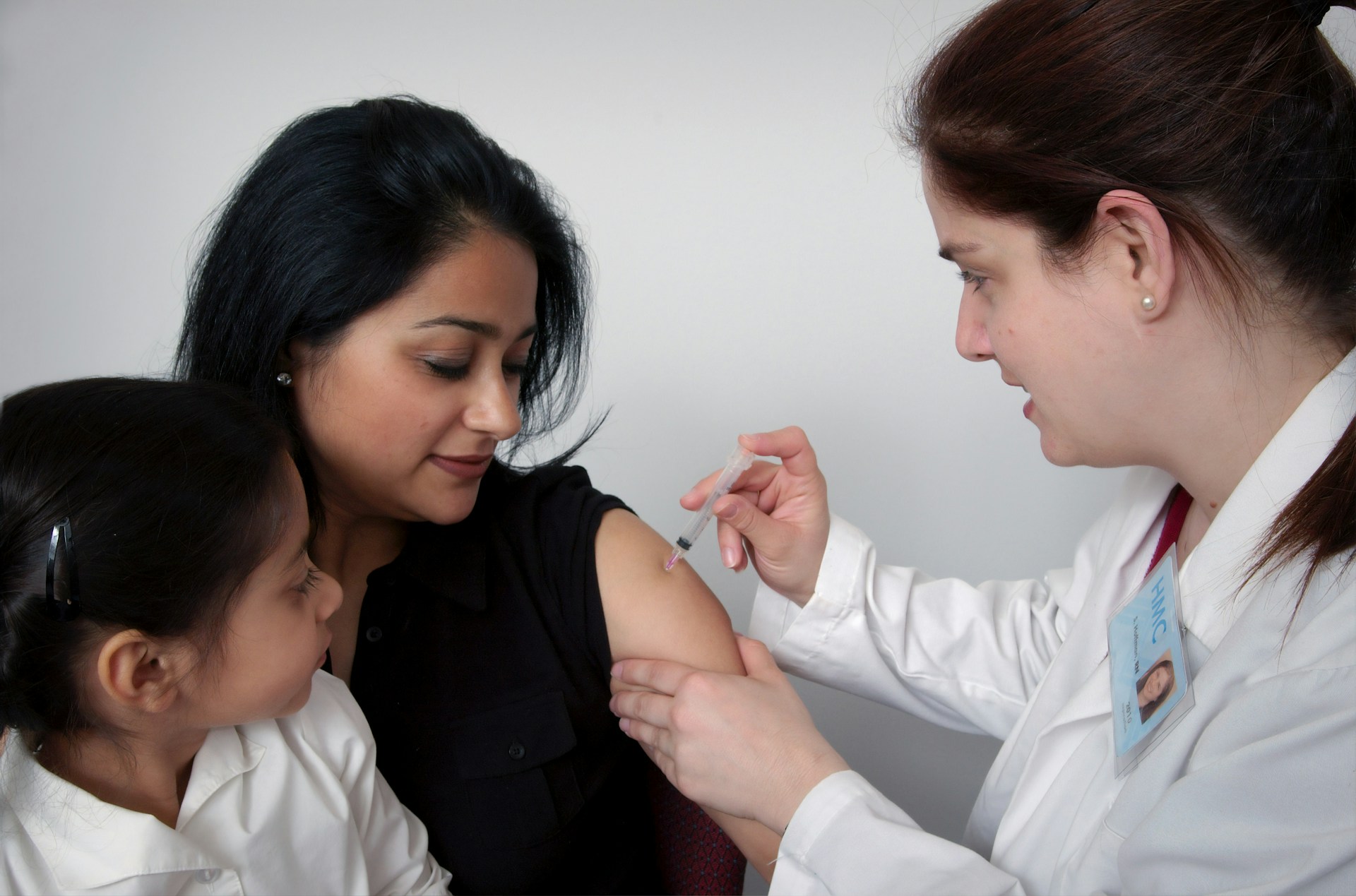 Public health nurse administering vaccine to woman with child observing.